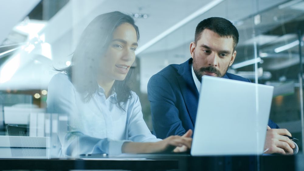 Female Office Worker Uses Smartphone while Sitting at Her Working Desk with Laptop on it.