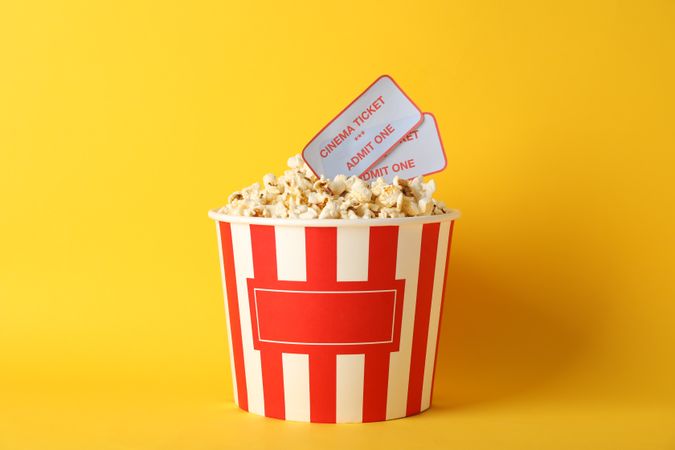 Striped bucket with popcorn and tickets on yellow background, copy space