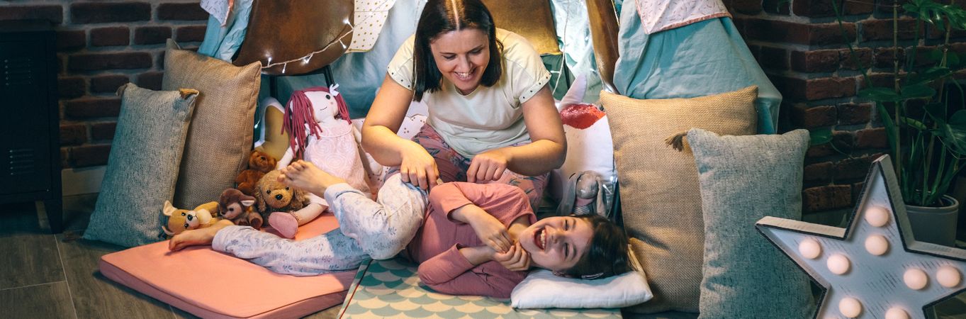 Banner of woman tickling to little girl while laughing lying over cozy mats in night pajama party