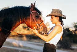 Young woman in hat walking with her horse in the countryside at sunset