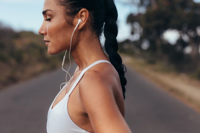 Woman in fitness gear standing on a road listening to music using earphones