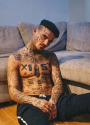 Shirtless Latino man covered in tattoos slumped against couch looking at camera