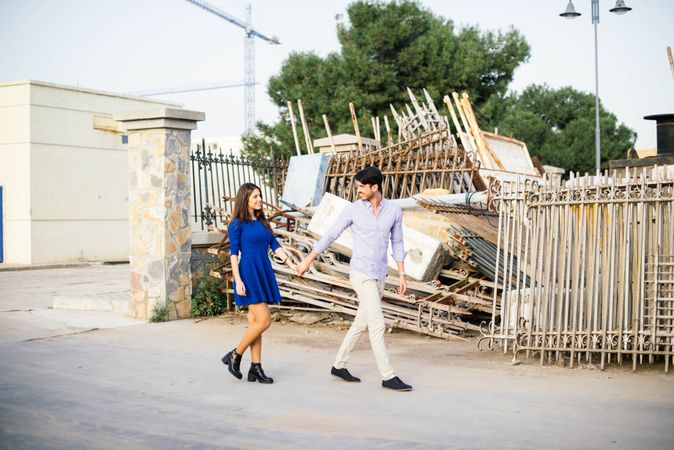 Loving couple walking holding hands along abandoned building with metal fence