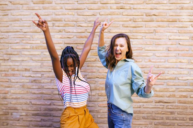 Two female friends with their arms up celebrating in front of a brick wall
