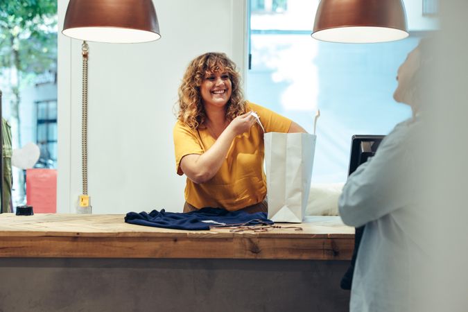 Store owner packing clothes in a paper bag looking at female customer and smiling