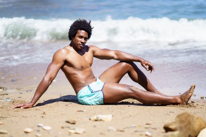 Man in swim trunks sitting down on beach near the water