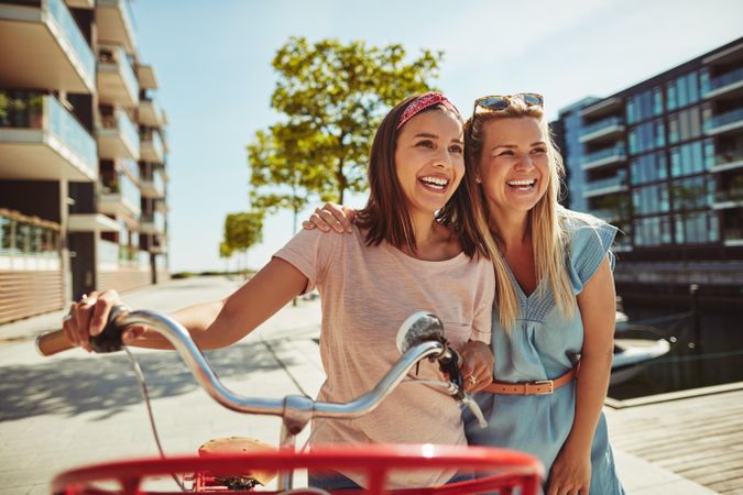 Two smiling female friends laughing while walking along a riverwalk with bike on sunny day, closeup