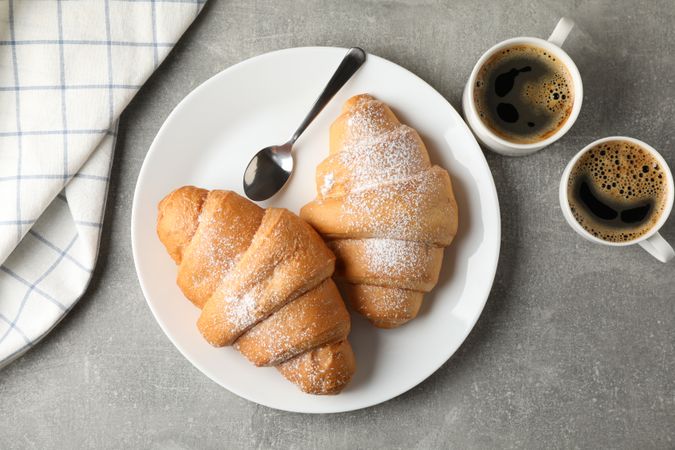 Plate with croissants, cups of coffee and towel on grey background, top view