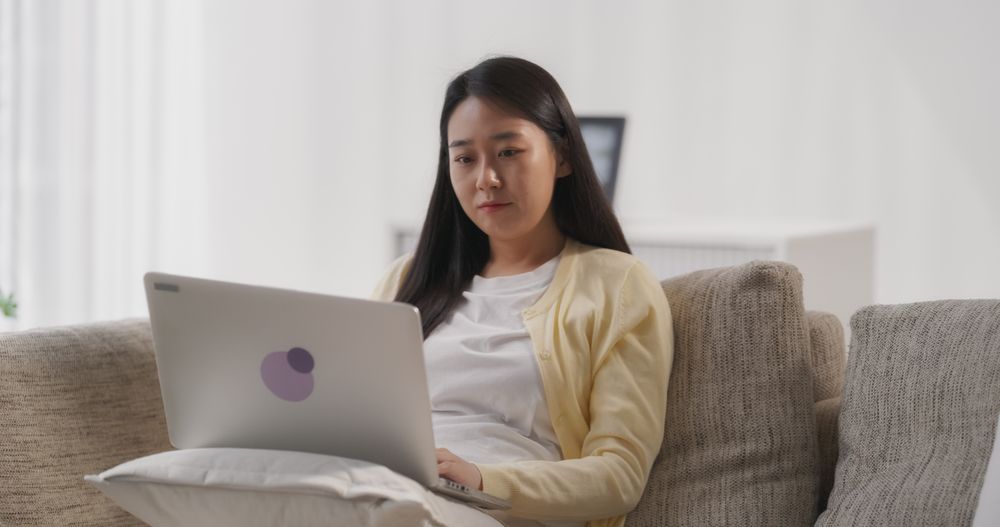 South Korean Woman Sitting on a Cozy Couch, Working on Laptop Computer on Her Project in a Stylish Bright Living Room