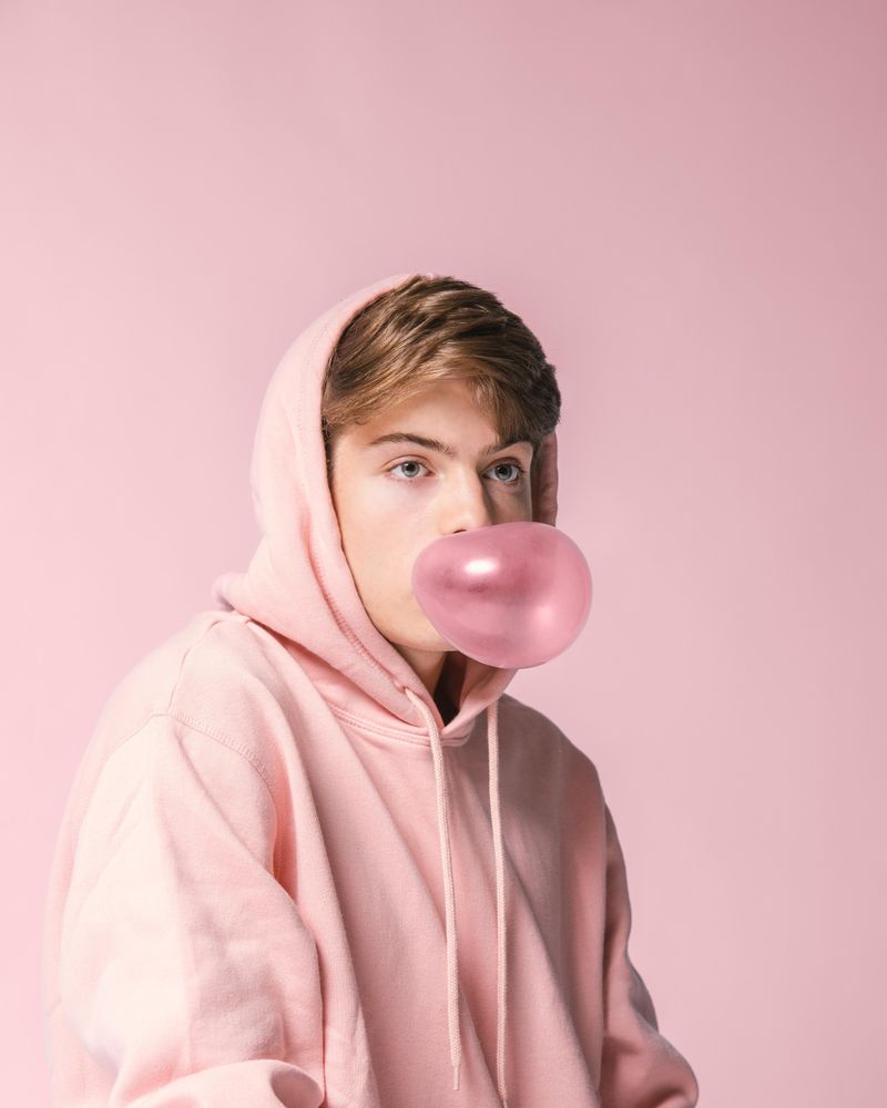 Teenage boy in a pink hoodie blowing bubble gum against pink background