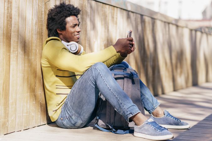Man sitting against wall outside and smiling while checking phone