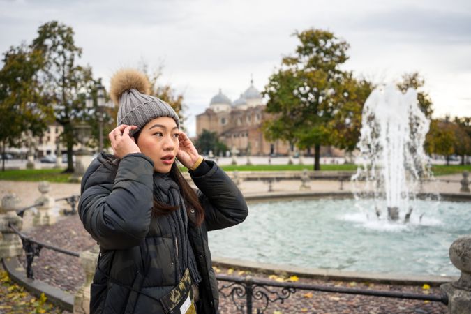Young female tourist in warm wear strolling at ancient park with beautiful buildings and fountain