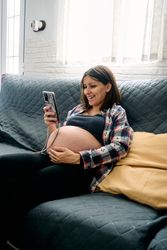 A young woman with a very advanced pregnancy is sitting on the couch looking at and talking on her cell phone.