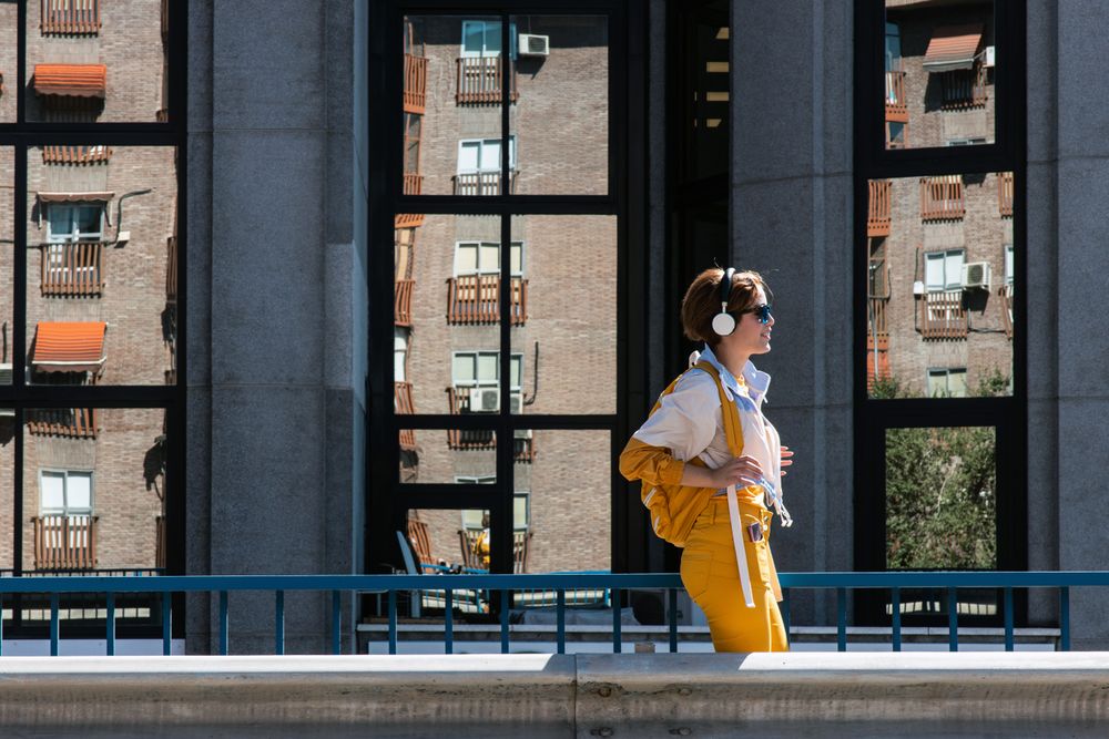 Stylish pensive young woman in sunglasses and headphones walking along road bridge in city