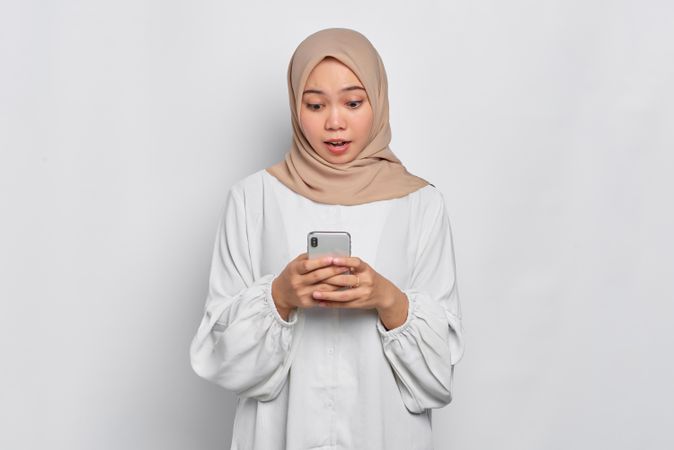 Amazed Asian female in headscarf looking down at cell phone