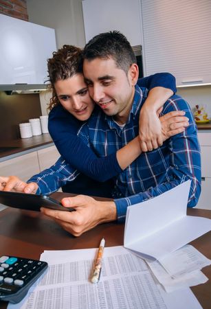 Woman holding man as they organize bills together in their kitchen, vertical