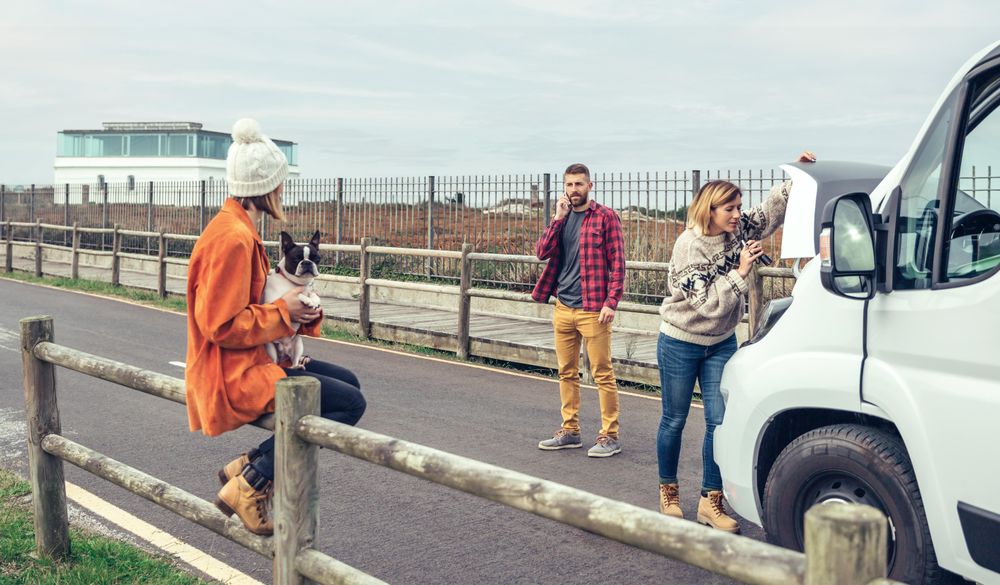 Woman in woolen sweater working on car engine while partner makes emergency call