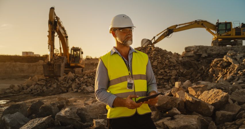 Male Civil Engineer Wearing Protective Goggles And Using Tablet On Construction Site On A Sunny Day. Man Inspecting Building Progress. Excavator Loading Materials Into Industrial Truck.