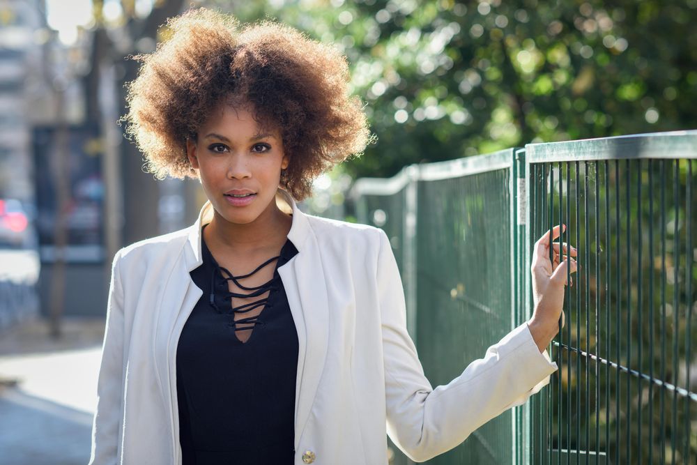 Female with afro looking at camera while standing on street next to green fence