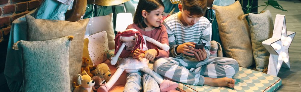 Banner of happy little girl embracing rag doll while looking boy playing game with phone sitting