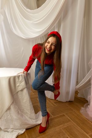Laughing teen girl in red adjusting shoes during photoshoot indoors.
