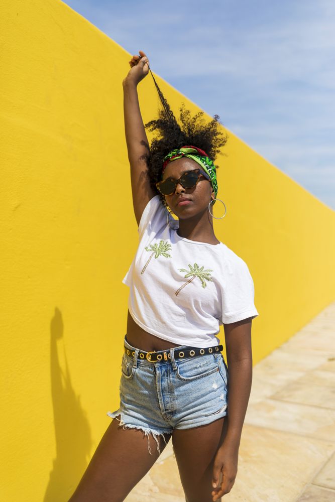 Young Black woman with sunglasses standing against yellow wall and playing with hair