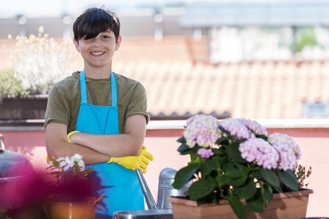 Young smiling teenager wearing gardening apron while standing with arms crossed on home terrace