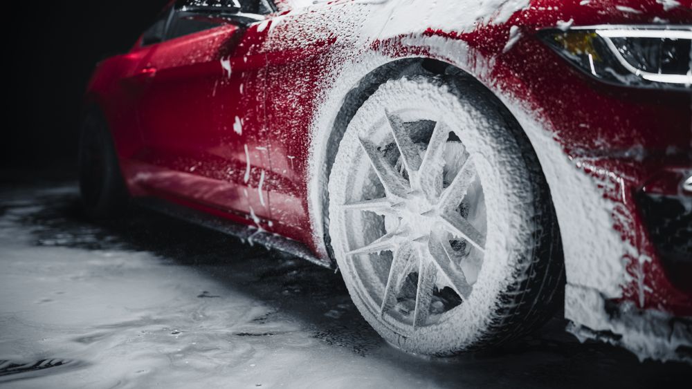 Professional Car Wash Worker Applying Smart Foam on a Front Wheel of a Red Sportscar with Retro Design at a Dealership Car Detailing Center.