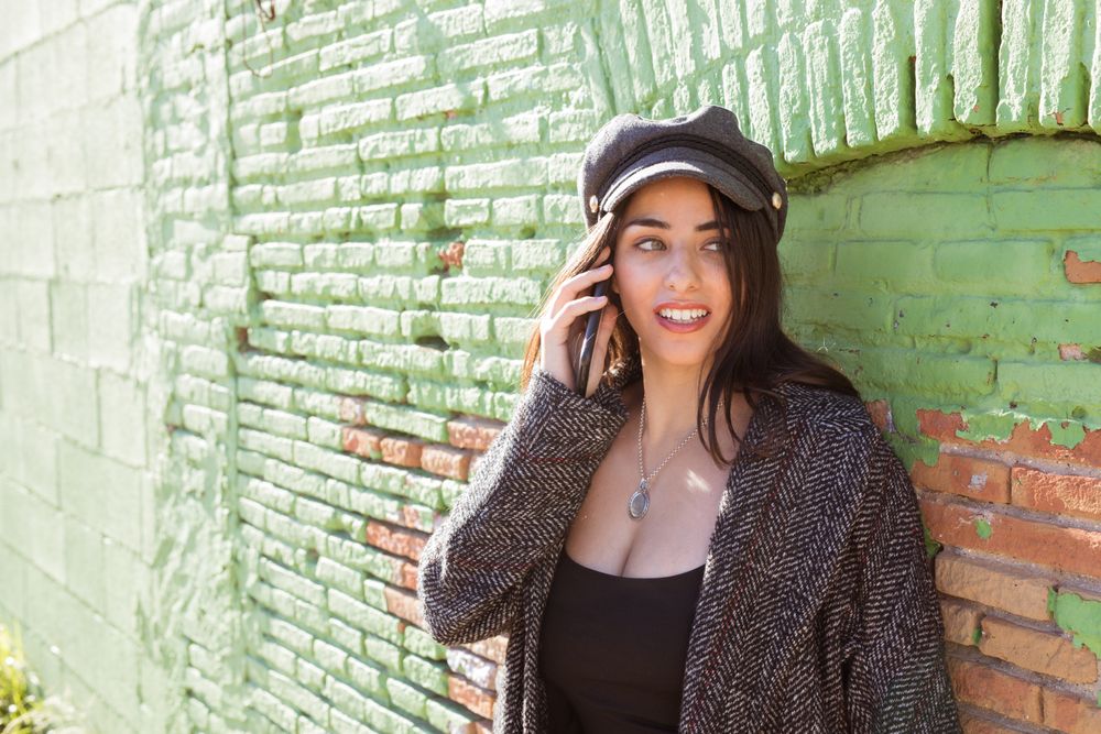 Charming Hispanic woman talking phone near wall