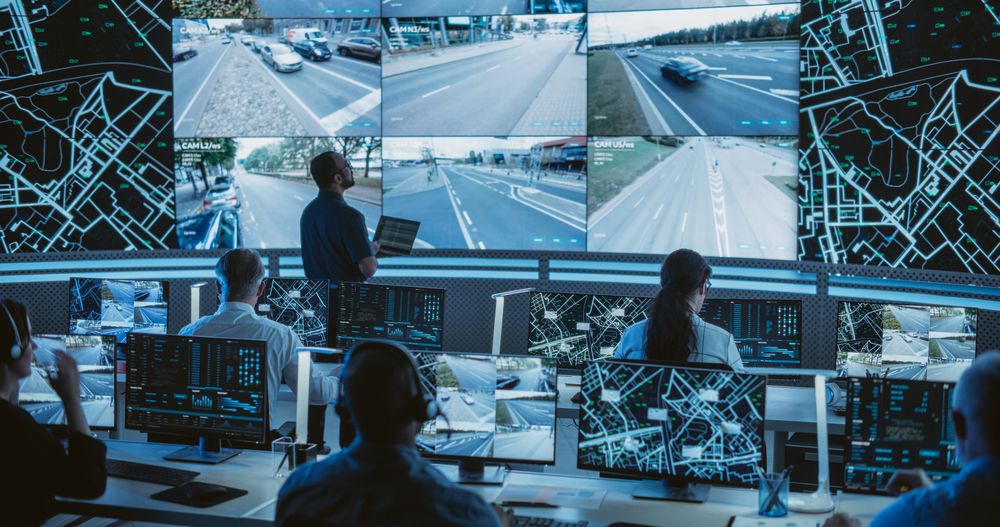 Male Software Engineer Working on Laptop Next to a Big Screen in a Modern Monitoring Office with Live CCTV Footage with Traffic Situation.