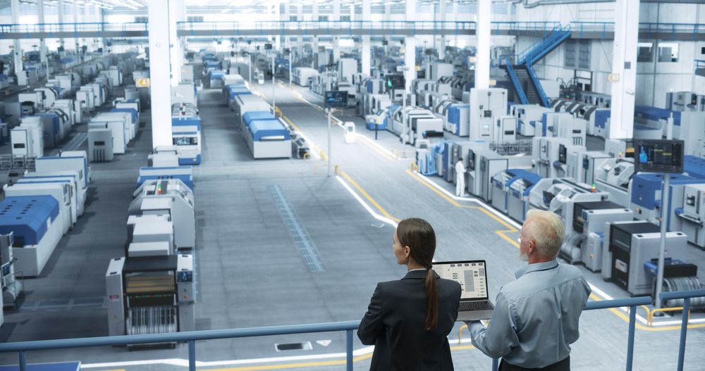 Male Engineer and Female Scientist Standing with Their Back, Using Laptop Computer and Talking in a Factory Facility with Equipment Producing Modern Technical Devices for Different Industries