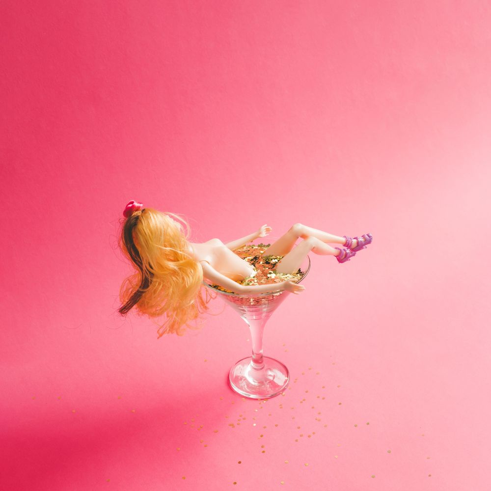 Doll bathing in martini glass full of gold glitter on pink background