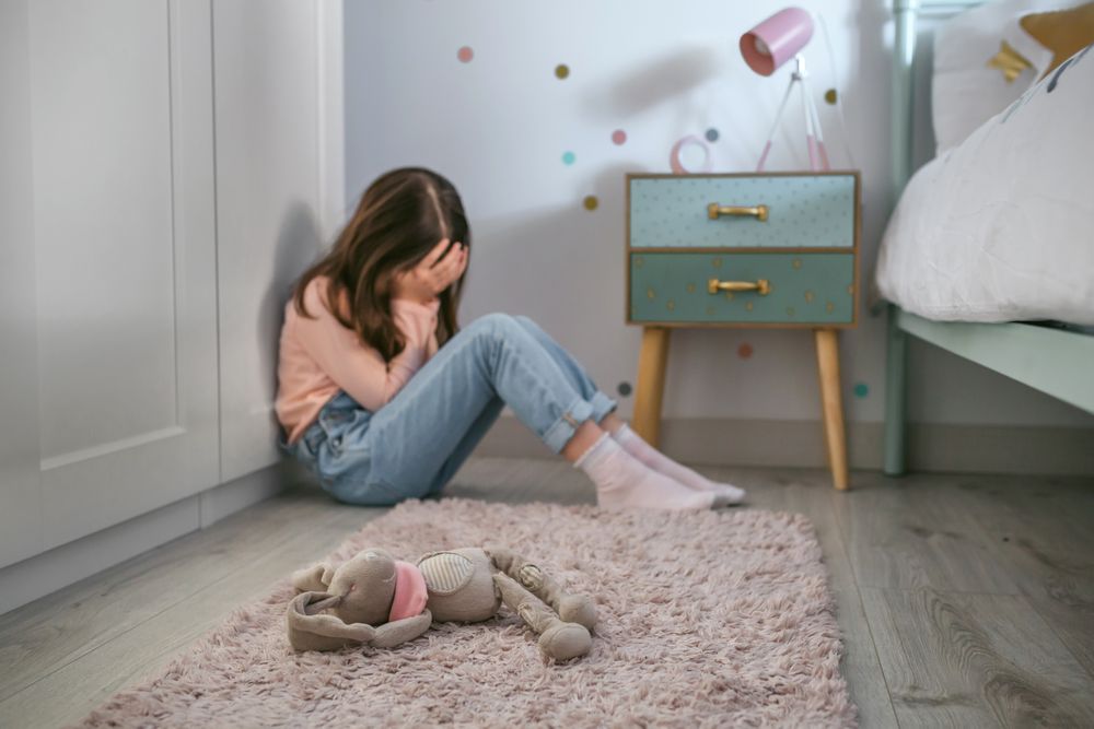Sad girl sitting on the floor of her bedroom with stuffed toy lying