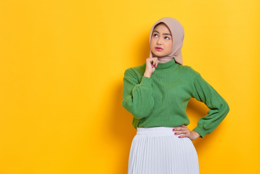 Woman in headscarf thinking with one hand on chin and the other on hip