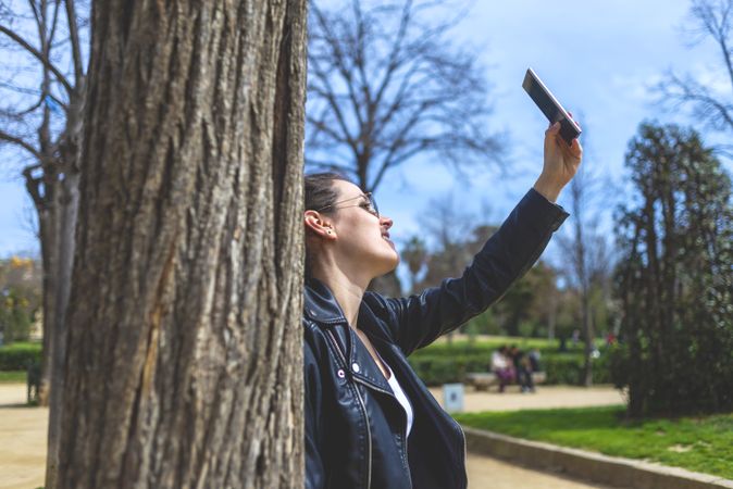 Side view of a young laughing hipster woman standing and leaning on a tree at park in sunny day while using a mobile phone to take a selfie