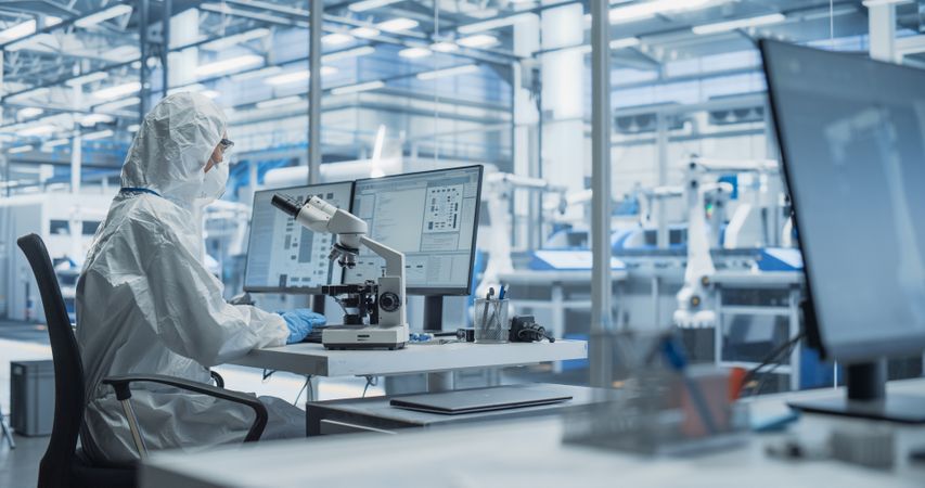 Modern Factory: Industrial Engineer Working on Desktop Computer in Clean Sterile Coveralls. Scientist Using a Microscope, Developing Advanced Solutions for High Tech Medical Vaccine and Gene Research