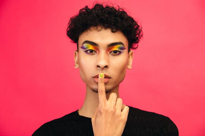 Male in rainbow eye make up gesturing silence, with finger on lips