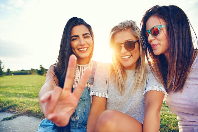 Happy female friends sitting in sunny field with one making the peace sign with hand