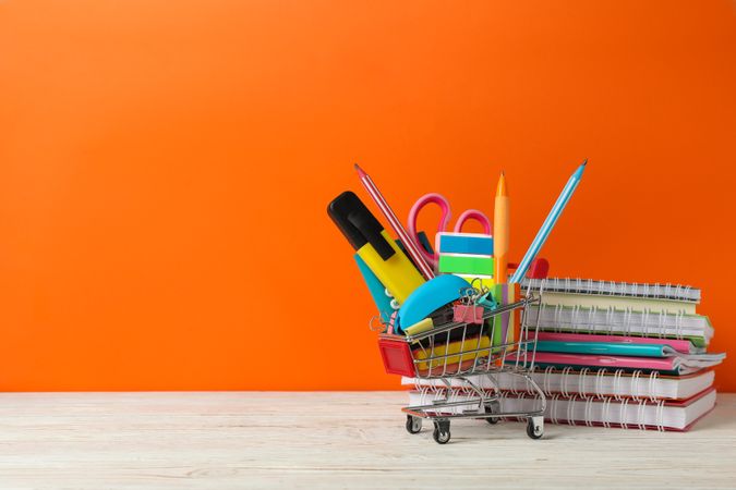 Shopping trolley with school supplies on orange background, space for text