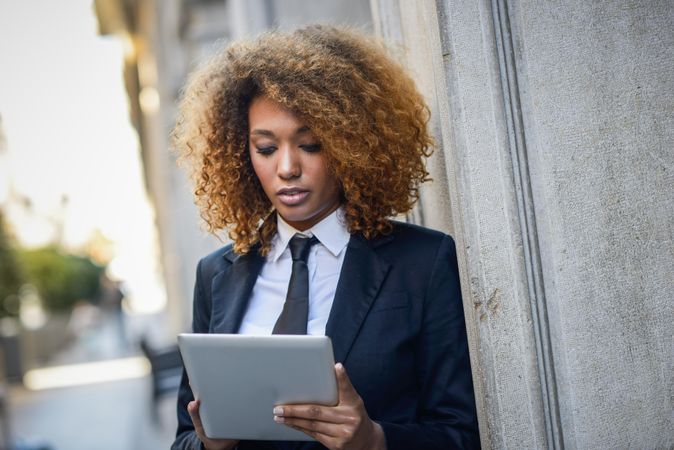 Serious female in suit standing outside looking down at her tablet