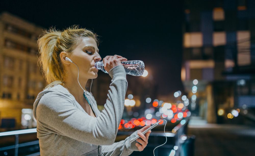 Portrait of tired female runner drinking water and looking exercise progress on tracker phone app