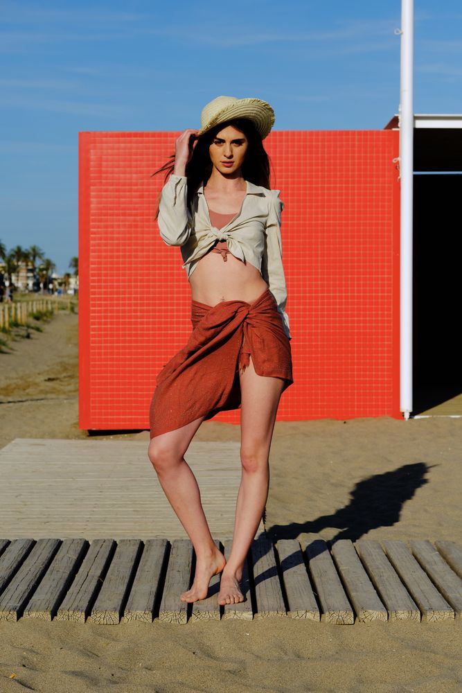 A long hair fashion model with a hat at the beach in front of a red wall