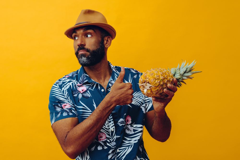 Male holding a pineapple to his chest with thumbs up sign in yellow studio