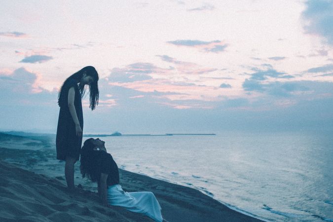 Side view of woman standing behind woman sitting on seashore at sunset