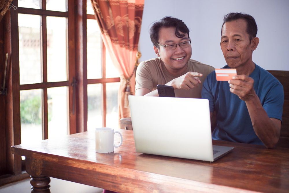 Happy adult son and older father pay online using laptop and credit card