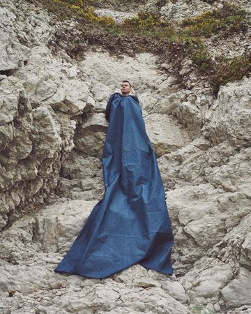 Young man draped in long blue fabric surrounded by a cliff