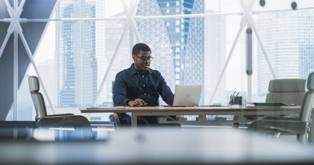Portrait of Successful Black Businessman Working on Laptop Computer in His Big City Corner Office.