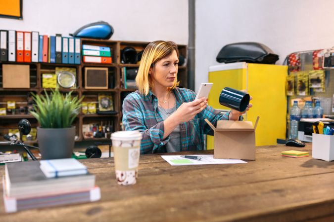 Young female entrepreneur taking photo with cell of a blue mug for client of her online store