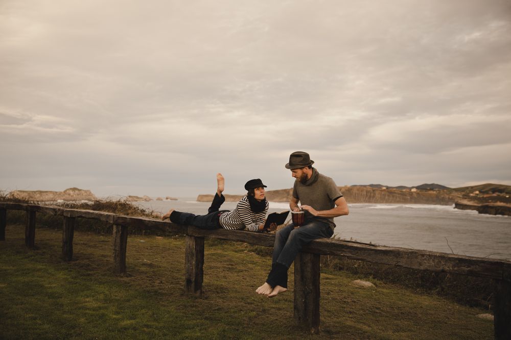 Man plying on drum near woman with notebook on bench on shore