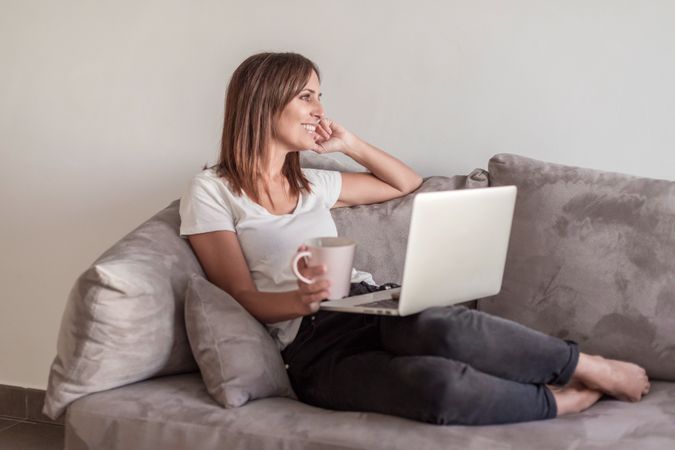 Happy woman on sofa with a cup of coffee working on her laptop in the morning